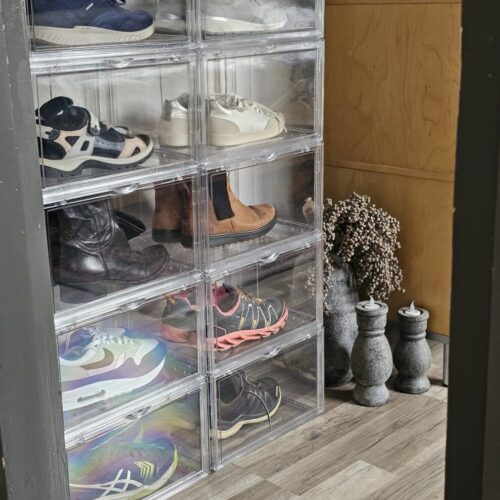 Tien transparante schoenendisplayboxen in twee kolommen gestapeld tot een grote opbergtoren, gevuld met diverse schoenen waaronder sneakers en laarzen, in een hal met houten vloer en decoratie.