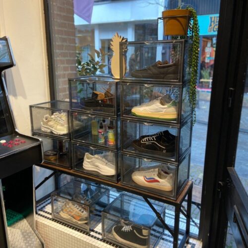Trapsgewijs gestapelde transparante sneakerdisplayboxen met diverse sneakers in een winkeletalage, met daarnaast een arcadekast en uitzicht op een winkelstraat.