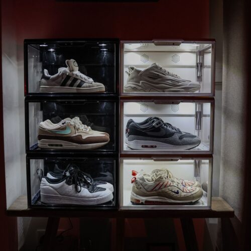 Zes verlichte zwarte en witte sneakerdisplayboxen met LED-verlichting in twee kolommen gestapeld, gevuld met diverse Nike Air Max- en Adidas-modellen, op een houten plank tegen een rode muur.