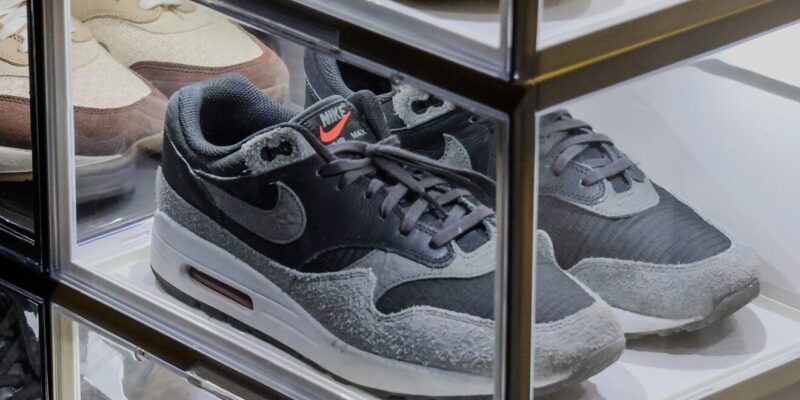 Close-up van een grijs-zwarte Nike Air Max 1 in een verlichte transparante displaybox, omringd door andere sneakers in gestapelde displayboxen.