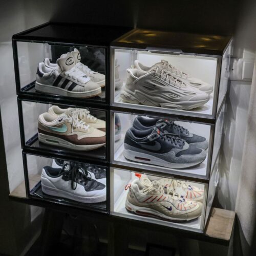 Zes verlichte witte en zwarte sneakerdisplayboxen met LED-verlichting in twee kolommen gestapeld, gevuld met diverse Nike Air Max- en Adidas-modellen, op een houten plank tegen een rode muur.