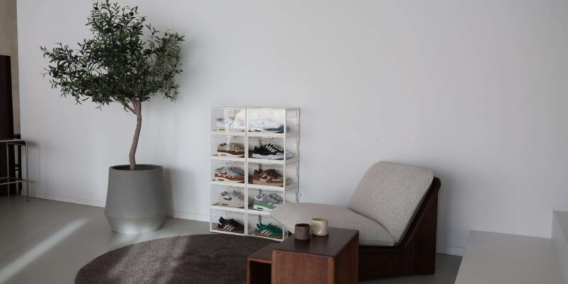 Witte sneakerdisplayboxen met LED in twee kolommen gestapeld tegen een witte muur, gevuld met Nike Air Max 1's en Adidas-modellen, naast een olijfboom in pot en een bouclé fauteuil in een licht, minimalistisch interieur.