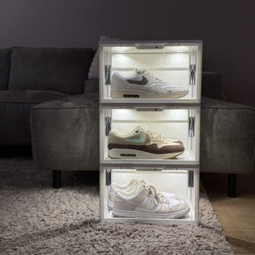 Drie verlichte witte sneakerdisplayboxen met LED-verlichting, op elkaar gestapeld met twee Nike Air Max 1's en een Nike Dunk Low, voor een grijze bank op een hoogpolig vloerkleed.